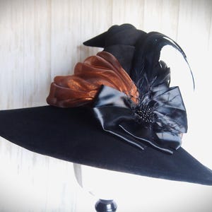 Pode incluir: Um chapéu preto com aba larga, adornado com tecido cor de cobre, penas pretas e um grande laço preto. O chapéu é exibido em um suporte branco, mostrando seu design elegante e detalhes.