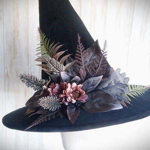 Pode incluir: Um chapéu de bruxa preto com aba larga, adornado com um arranjo de flores e folhagens artificiais. O arranjo floral inclui folhas e flores bordô escuro, malva e verdes, criando uma estética gótica e outonal.