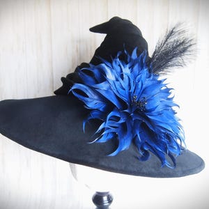 Pode incluir: Um chapéu de bruxa preto com aba larga, adornado com uma grande flor de penas azul vibrante e uma pena preta. O chapéu é exibido em um suporte branco, mostrando seu design elegante.