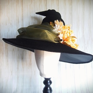Pode incluir: Um chapéu de bruxa preto com aba larga, adornado com um aglomerado de flores laranja e brancas e tecido verde-oliva. O chapéu é exibido em uma cabeça de manequim contra um fundo de madeira clara.