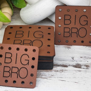 Peut inclure: Sous-verres carrés en cuir marron avec les mots "BIG BRO" gravés en noir. Chaque dessous de verre comporte une série de petits trous sur les bords. Une pile de dessous de verre est visible, ainsi que des dessous de verre individuels.