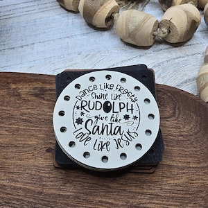 May include: A stack of square coasters with a white top coaster featuring the text "Dance like Frosty, Shine like Rudolph, Give like Santa, Love like Jesus." The coasters are black and white and are on a wooden surface.