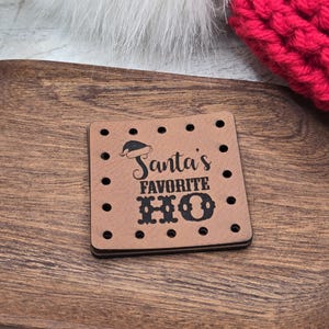 May include: A square tan leather patch with pre-punched holes along the edges. The patch features a black engraved graphic of a Santa hat above the text Santa's FAVORITE HO, with the word HO displayed in a large, decorative, bold black font.