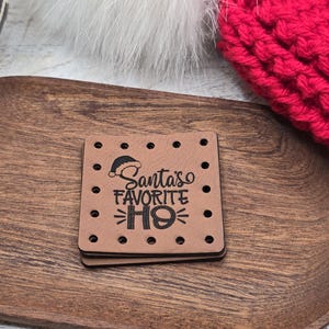 May include: A square brown leather tag with rounded corners and pre-punched holes along the edges. The tag features a black printed graphic of a Santa hat and the text Santa's FAVORITE HO, with the letter O stylized as a tire tread pattern.