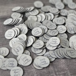 May include: A close-up of a pile of white round labels with the text "Little Lady Crochets" printed on them.