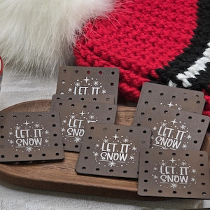 May include: A set of brown square coasters with the text "LET IT SNOW" in white, arranged on a wooden tray. The coasters have small holes around the edges. A red and black knitted hat is in the background.