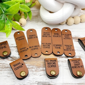 Peut inclure: Neuf organisateurs de cordon en cuir brun avec différentes citations sur le thème du café. Les citations incluent "Coffee Time", "But First, Coffee", "No Talkie Before Coffee", "Shhh... Coffee", et "Shhh... The Coffee Hasn't Kicked In Yet".