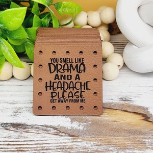 May include: A stack of brown rectangular tags with rounded corners and small holes. The tags have the text "YOU SMELL LIKE DRAMA AND A HEADACHE PLEASE GET AWAY FROM ME" in black. The background includes greenery and white wooden beads.