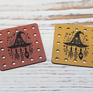 May include: Two square leather patches, one rust-colored and one mustard-yellow, each featuring a black laser-engraved design of a witch hat surrounded by flowers, herbs, and a crescent moon. Both patches have pre-punched holes along the edges.