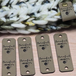 Peut inclure: Gros plan sur plusieurs étiquettes rectangulaires beiges avec le texte "Yarnful HEARTS" imprimé en noir. Les étiquettes comportent un petit cœur et un graphique de pelote de laine. Un article tricoté est en arrière-plan. Des rivets en laiton sont également visibles.