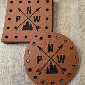 May include: A stack of square brown leather tags and a single circular brown leather tag, each featuring pre-punched holes along the edges. The tags are printed with a black graphic of two crossed arrows, the letters P, N, and W, and pine trees.