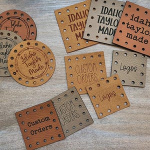 May include: A collection of circular and square leather tags in shades of tan and gray, each featuring pre-punched holes along the edges. The tags are laser-engraved with text including Idaho Taylor Made, Custom Orders, and Logos in various fonts.