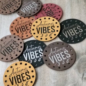 May include: A collection of circular leather tags in various colors including tan, brown, black, and maroon. Each tag features a series of pre-punched holes around the perimeter and the text "Autumn VIBES" engraved in the center in a script and block font.