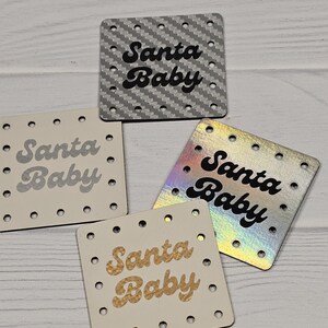 May include: Four square tags with rounded corners and pre-punched holes along the edges. Each tag features the text "Santa Baby" in a bold, retro-style font. The tags include a silver carbon fiber texture, a matte cream, a gold glitter, and a holographic finish.