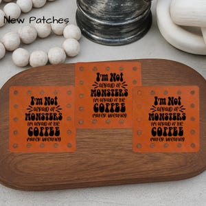 May include: Three orange square patches with the text "I'm Not afraid of MONSTERS I'm afraid of the COFFEE maker breaking" in black. The patches are on a wooden tray with decorative white spheres and a metal container.