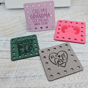 May include: Four square leatherette patches with decorative text and designs. One is lavender with "CALL MY GRANDMA SHE KNOWS WHAT TO DO". Another is pink with baby footprints. A green patch says "Santa's cutest Elf". The last is grey with "it's a Girl!"