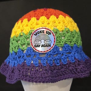 May include: A crochet bucket hat featuring horizontal stripes in red, orange, yellow, green, blue, and purple. A circular pin is attached to the front, displaying a rainbow graphic and the text WOKE UP GAY AGAIN in black capital letters.