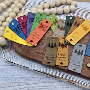 May include: A selection of colourful tags with the text "MADE WITH LOVE" and "Russell Sprouts Knits". The tags are in various colours, including yellow, blue, red, and green, and feature a tree design. They are arranged on a wooden surface.