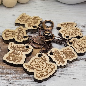 May include: A set of eight wooden keychain charms featuring Highland Cow designs. Each charm is laser-cut and engraved with a different Highland Cow pose. The charms are attached to a metal ring with a lobster clasp.
