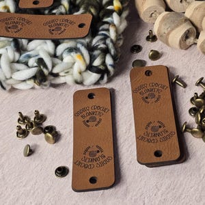 May include: Close-up of leather tags with the text "Sadie's Crochet Blankets" stamped on them. The tags are brown with rounded corners and holes for attaching to blankets. Also visible are metal rivets, wooden spools, and a crocheted blanket.