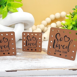 May include: Three brown leather squares with the words "So Loved" and "John 3:16" in black script. Each square has a heart symbol and small dotted holes around the edges. The squares are arranged on a white wooden surface.
