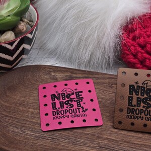 May include: Two square leather patches with the text "NICE LIST DROPOUT (GOPPY SANTA)". One patch is pink, the other is brown. Both have small holes around the edges. The patches are on a wooden surface.