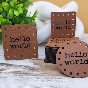 May include: Brown leather tags with the words "hello world" printed in black. The tags are square and round, with evenly spaced holes around the edges. A stack of tags is visible, along with a few individual tags.
