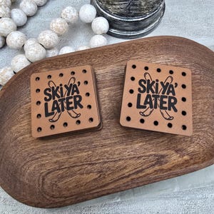 May include: Two square brown coasters with the text "Ski Ya Later" in black, featuring crossed skis. The coasters are on a wooden tray. Small black dots surround the text. A stack of coasters is visible.