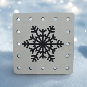 May include: A square, light-colored wooden block with a black snowflake design in the center. The block has evenly spaced holes around the edges. The background is a blurred winter scene with white and blue tones.