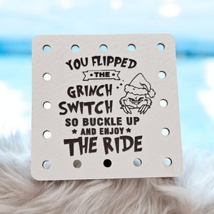 May include: A square, off-white sign with the text "YOU FLIPPED THE GRINCH SWITCH SO BUCKLE UP AND ENJOY THE RIDE" in black. The Grinch is illustrated wearing a Santa hat. The sign has several evenly spaced holes around the edges.