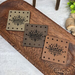 May include: Three square leather tags with the word "Winter Adventures" and a snowflake design. The tags are in shades of brown and tan, each with small holes around the edges, displayed on a wooden tray.