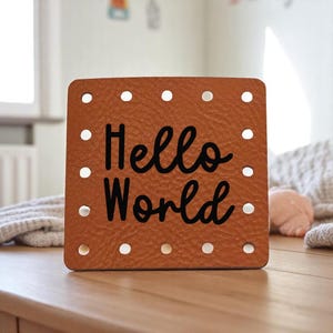 May include: A square, brown leather sign with the words "Hello World" in black script. The sign has evenly spaced white dots around the edges, and is sitting on a wooden surface.