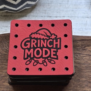 May include: A stack of square red coasters featuring the text "GRINCH MODE" in black, a Santa hat, and fairy lights design. Each coaster has small holes around the edges. The coasters are on a wooden surface.