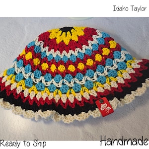 May include: A crocheted bucket hat featuring a vibrant granny square pattern in red, yellow, blue, and cream yarn. The hat includes a scalloped cream edge and a small red rectangular tag with a white mushroom graphic and the text Idaho Taylor Made.