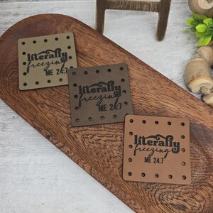 May include: Three square leather patches in shades of brown are displayed on a wooden tray. Each patch has the phrase "literally freezing ME 24:7" printed in black. The patches have small holes around the edges.
