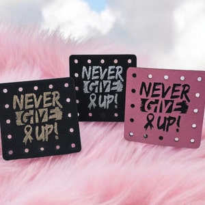 May include: Three square coasters with the text "NEVER GIVE UP!" in silver, black, and pink. Each coaster has a dotted border and a ribbon graphic. The coasters are on a pink, fluffy surface.