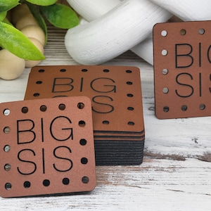 Peut inclure: Des étiquettes carrées marron avec l'inscription "BIG SIS" en noir. Chaque étiquette est percée de petits trous espacés sur les bords. Une pile d'étiquettes et quelques étiquettes individuelles sont visibles, suggérant un ensemble ou une collection.