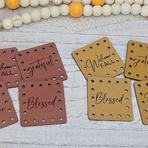 May include: Eight square faux leather patches with pre-punched holes along the edges. Four patches are dark brown and four are tan. The text printed in black includes the phrases Welcome FALL, grateful, Blessed, and Sweater Weather.
