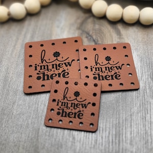 Peut inclure: Trois étiquettes carrées en cuir marron avec le texte "hi i'm new here" en écriture noire. Chaque étiquette comporte une série de trous espacés régulièrement sur les bords. Un motif floral est au-dessus du "i".