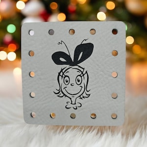 May include: A white square-shaped coaster with a black and white illustration of a girl with a bow on her head. The coaster has multiple holes around the edges.