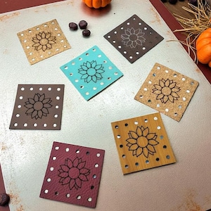May include: A collection of square coasters in various colors, including brown, teal, and burgundy. Each coaster features a laser-cut sunflower design and small circular perforations around the edges. The coasters are arranged on a neutral surface, with pumpkins and decorative elements in the background.