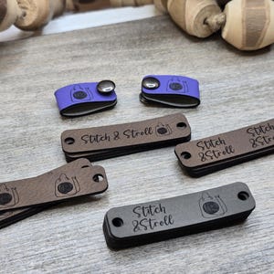 May include: A selection of leather tags and snap closures in different colours. The tags are brown and grey, with the text "Stitch & Stroll" and a shopping bag graphic. Two purple snap closures are also visible.