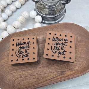 May include: Two square brown coasters with the text "When in doubt, Ski it out" in black. Each coaster has small, evenly spaced holes around the edges. The coasters are stacked on a wooden tray.