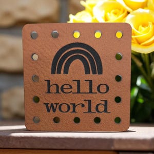 Peut inclure: Une étiquette carrée en cuir beige aux coins arrondis avec un motif arc-en-ciel noir imprimé au-dessus du texte hello world. L'étiquette présente une série de trous circulaires régulièrement espacés sur les quatre bords pour la couture.