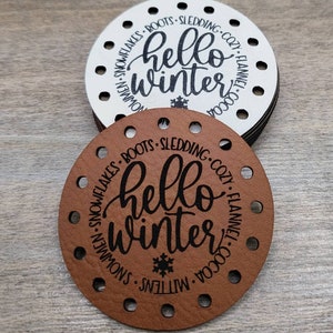 May include: A stack of circular tags with pre-punched holes around the perimeter. The top tag is brown, and the one beneath is white. Both feature the black text "hello winter" in the center, surrounded by words like "boots," "sledding," and "cocoa."