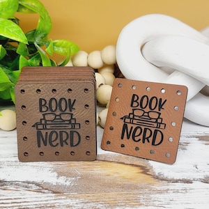 May include: Two brown leather patches with a black design that says "Book Nerd" with a pair of glasses and a stack of books. The patches have a dotted pattern around the edges.