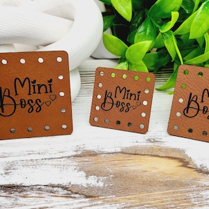 May include: Three brown leather tags with the words "Mini Boss" in black script, each with small silver rivets around the edges. The tags are square and vary in size, with a heart symbol next to the word "Boss".