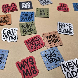 May include: A collection of colorful, square-shaped patches, each featuring the name of a U.S. state in a unique font. The patches are in various colors, including red, blue, green, and brown, with white lettering. Some patches have decorative holes around the edges.