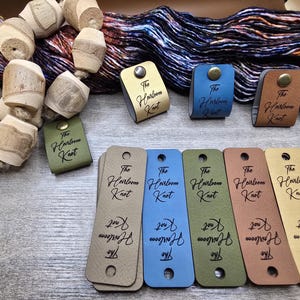 Peut inclure: Un ensemble d'étiquettes en cuir et de fil. Les étiquettes sont de différentes couleurs, dont le beige, le bleu et le vert, avec le texte "The Heirloom Knot" imprimé dessus. Des perles en bois et une pelote de fil sont également présentes.