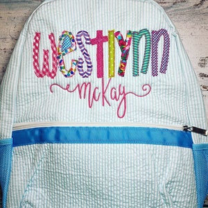 May include: A light blue and white striped backpack with the name "Westyn McKay" embroidered in colorful, patterned letters. The backpack has a blue zipper, trim, and mesh side pockets. The bag is designed for carrying school supplies.